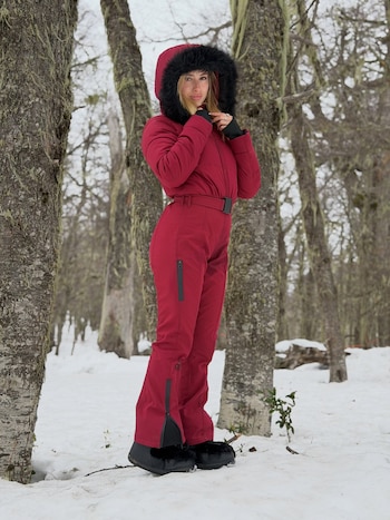 Una mujer viste un traje térmico rojo con capucha de piel sintética negra sobre la nieve rodeada de árboles