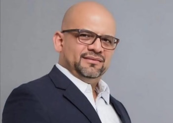 Primer plano de un hombre calvo con barba y gafas, vistiendo un traje oscuro y camisa blanca, mirando directamente a la cámara sobre un fondo gris liso