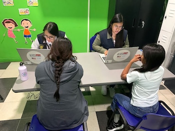 Dos mujeres con chalecos y laptops frente a dos jóvenes en un espacio con pared verde decorada, mesas grises y sillas azules. Una mujer y una joven de espaldas