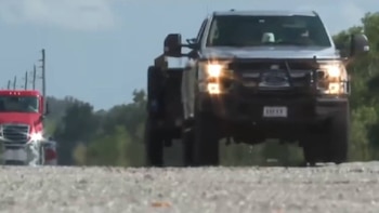 Un vehículo Ford oscuro tipo SUV o todoterreno se ve de frente, con las luces encendidas, circulando por un camino de grava bajo un cielo claro