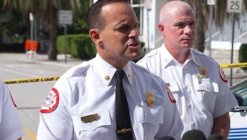 Robert Hevia viste uniforme blanco de bomberos y corbata negra mientras habla ante micrófonos junto a otro oficial en la vía pública