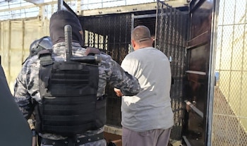 Oficial con uniforme de camuflaje, chaleco y pasamontañas escolta a hombre rapado de camiseta gris. Fondo con rejas metálicas