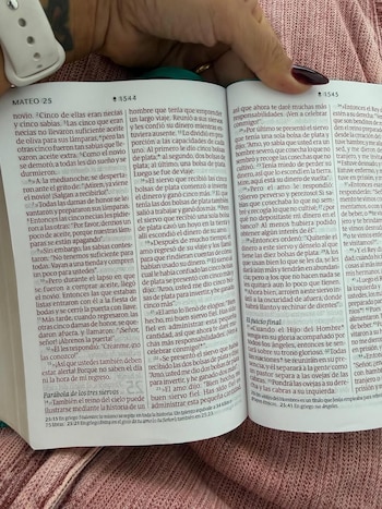 Primer plano de una mano con manicura roja oscura sosteniendo una Biblia abierta en la página que muestra el capítulo 25 de Mateo