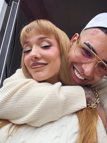 Una mujer rubia con flequillo, piercings faciales y maquillaje abraza a un hombre con gafas, piercings faciales, tatuajes y un gorro. Ambos sonríen