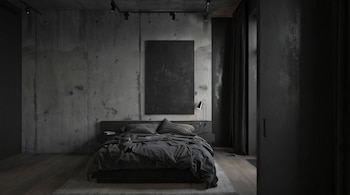 Dormitorio moderno con paredes de hormigón gris, una cama doble grande con ropa de cama gris oscuro y almohadas, un cuadro abstracto negro vertical y una lámpara de mesa a la derecha.