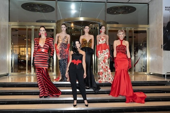 Diseñadora y cinco modelos posan frente a una puerta dorada. Modelos visten vestidos de gala, rojos, estampados y negros con detalles dorados y rojos
