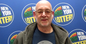 Primer plano de un hombre calvo con gafas, usando un auricular, y vestido con una chaqueta gris, frente a un fondo azul con logos de la Lotería de Nueva York