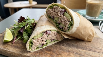 Dos mitades de wrap de atún con lechuga, cebolla morada y pepino, envueltas en papel, junto a una ensalada verde y rodaja de lima en tabla de madera.