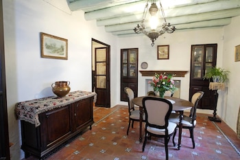 Una sala con paredes blancas, techo de vigas azules, un candelabro, mesa redonda con sillas, un aparador, cuadros, armarios y plantas