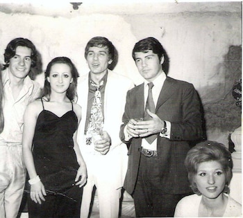 Cinco personas en blanco y negro: cuatro de pie y una sentada. Todos llevan ropa formal o semi-formal de estilo vintage, incluyendo trajes, vestidos y camisas
