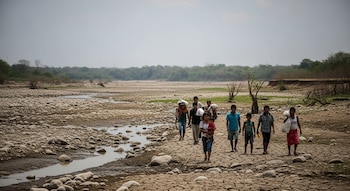 Grupo de personas, con mujeres, hombres y niños, camina por un río seco con lecho de piedras. Algunas llevan bultos. Vegetación y árboles al fondo.