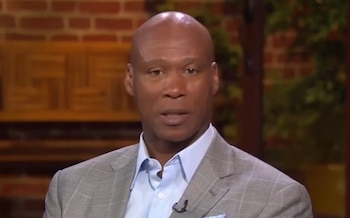 Retrato de Byron Scott con chaqueta gris de cuadros y camisa azul claro en un estudio con fondo de ladrillos