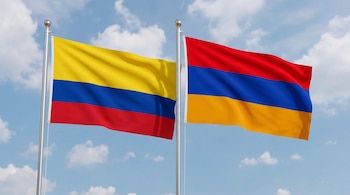 Dos banderas ondean, la de Colombia (amarillo, azul, rojo) y la de Armenia (rojo, azul, naranja), contra un cielo azul claro con nubes blancas.