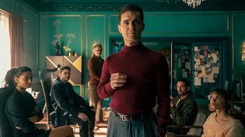 Pedro Alonso, con jersey rojo y pantalón gris, mira a cámara, rodeado de otras personas en una habitación con paredes aguamarina y molduras doradas