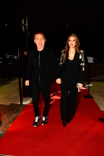 Hombre y mujer tomados de la mano en alfombra roja. Él con traje negro y zapatillas, ella con traje negro, bordados en mangas, tacones y teléfono móvil
