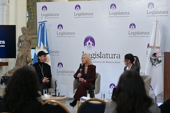 Panel de tres personas en un evento de la Legislatura de Buenos Aires, sentadas frente a un fondo con logos y la bandera argentina. El público está en primer plano desenfocado