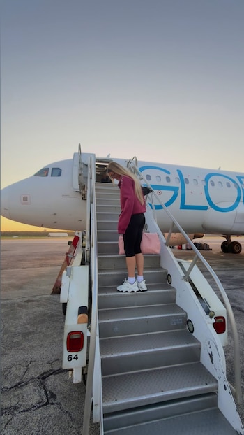 Una mujer rubia con sudadera rosa y leggings negros sube las escaleras de un avión blanco con letras azules en la pista de un aeropuerto