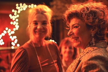 Una mujer de cabello castaño rojizo rizado y grandes pendientes sonríe y mira hacia la izquierda, con otra mujer rubia sonriente detrás