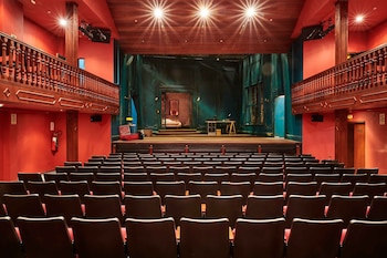 Sala de teatro con butacas oscuras, paredes rojas, balcones laterales de madera y un escenario iluminado con decorado de interiores