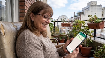 Mujer sentada en un sillón en un balcón con plantas mientras sostiene un teléfono móvil que exhibe un mensaje de texto en la pantalla.