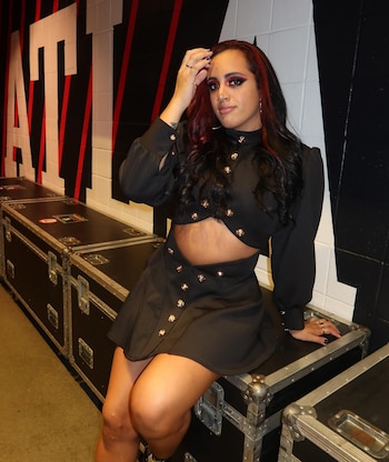 Simone Johnson sentada sobre estuches negros, con un top corto y falda negra, cabello oscuro con mechas rojas y maquillaje intenso