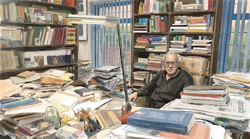 Dibujo en acuarela de un hombre mayor con gafas sentado en un escritorio abarrotado de libros y papeles. Detrás, estanterías repletas y una lámpara de escritorio.