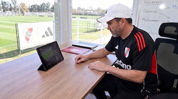 Un hombre con gorra blanca y camiseta negra con escudo de River Plate, sentado frente a una tableta y una pizarra con notas, con un campo de fútbol de fondo.