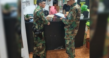 Dos hombres en uniformes militares de camuflaje se encuentran frente a un mostrador. Uno escribe en un portapapeles azul, el otro observa. Hay plantas sobre el mostrador
