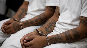 Brazos y torsos de dos personas sentadas con camisas blancas, brazos esposados con esposas de metal y antebrazos con tatuajes.