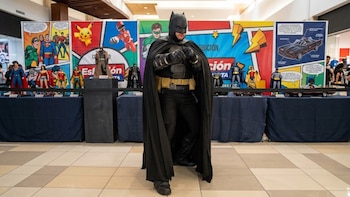 Un hombre disfrazado de Batman posa con los puños en alto frente a mesas con figuras de coleccionista y carteles de historietas
