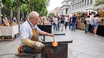 Un hombre con delantal y guantes moldea vidrio incandescente con varas de metal frente a puestos de artesanía y peatones en una calle.