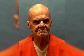 Retrato de un hombre de edad avanzada con uniforme de prisión naranja y camiseta blanca frente a un fondo neutro