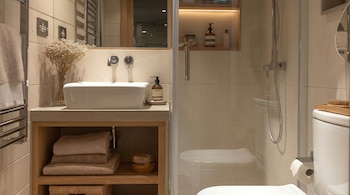 Vista de un baño moderno con lavabo rectangular, grifo de pared, estantes de madera, toallas, flores secas, ducha de cristal, inodoro y detalles decorativos.