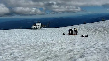 Un helicóptero blanco está posado en una ladera nevada, con un grupo de personas y equipo de rescate cerca, y dos figuras tumbadas sobre la nieve