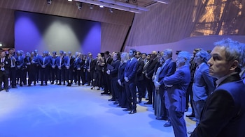 Un grupo de personas vestidas con trajes oscuros permanece de pie en un amplio salón iluminado con luces azules frente a un escenario