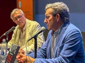 Dos hombres senior en un panel. Uno, con chaqueta azul, habla a un micrófono, mientras el otro, con chaqueta beige, escucha. Un libro y agua sobre la mesa