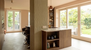 Interior moderno de cocina y comedor con columna de azulejos verdes, isla con encimera de mármol y estantes de madera, grandes ventanales a un jardín.