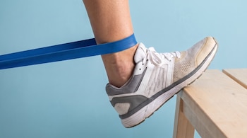 Pierna de persona con zapatilla blanca y gris y banda de resistencia azul alrededor del tobillo, apoyada sobre un banco de madera, fondo celeste.