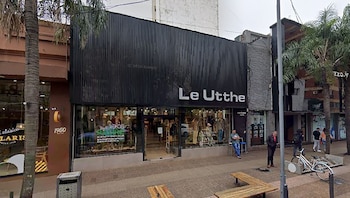 Vista exterior de la tienda Le Utthe en Posadas, Argentina. Fachada negra moderna con grandes ventanales, mostrando maniquíes. En la acera, personas, bancos y árboles