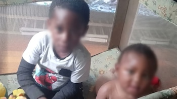 Dos niños con los rostros difuminados se encuentran dentro de un corralito de red con una colchoneta estampada y juguetes.