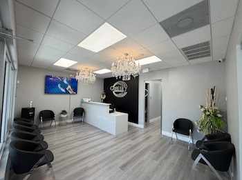 Sala de recepción con un mostrador blanco, nueve sillas negras, dos candelabros de cristal, televisor en la pared, paredes grises y piso de madera clara