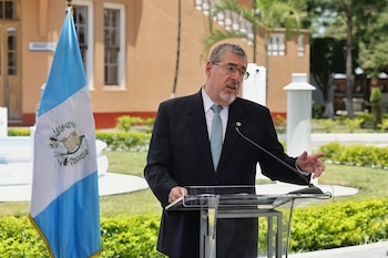 Hombre canoso con traje oscuro y corbata azul claro habla en un podio transparente, con una bandera de Guatemala a la izquierda y un edificio ocre al fondo