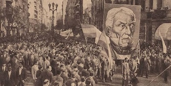 Fotografía en blanco y negro de una multitud en una calle, con personas que llevan banderas y un cartel gigante con un retrato