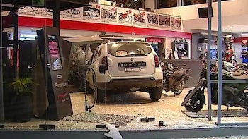Un SUV blanco está incrustado en la vidriera rota de una tienda de motocicletas, con fragmentos de vidrio cubriendo el suelo. Motocicletas y cascos son visibles