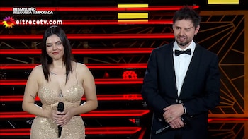 Una mujer con vestido dorado brillante y un hombre con esmoquin negro y pajarita sostienen micrófonos frente a un fondo rojo