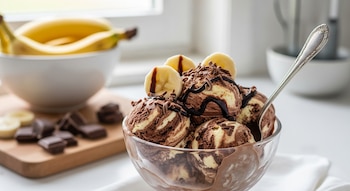 Bol de cristal con helado de chocolate y plátano, cubierto con sirope, virutas y rodajas de plátano. Al fondo, plátanos y trozos de chocolate.