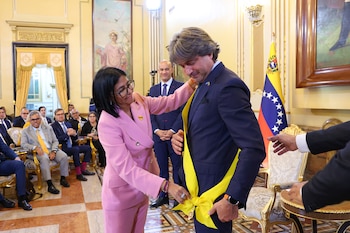 Una mujer con traje rosado coloca una banda amarilla a un hombre con traje oscuro frente a la bandera de Venezuela y varias personas