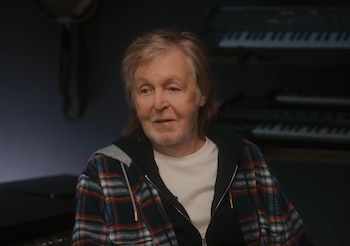 Primer plano de Paul McCartney con cabello y barba canosos, vistiendo una camisa a cuadros y sudadera. Mira pensativo, con teclados musicales borrosos al fondo
