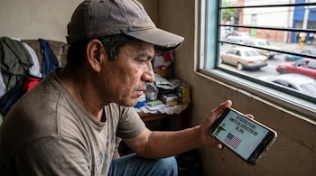 Hombre salvadoreño con gorra y camiseta mira un teléfono con pantalla rota. El móvil muestra el texto 'ESTATUS LEGAL: ÚNETE AL EJÉRCITO DE EE. UU.' con la bandera de EE. UU.