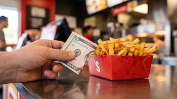 Una mano sostiene un billete de cinco dólares junto a una caja roja con papas fritas sazonadas sobre un mostrador metálico.